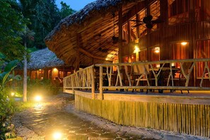 Un bâtiment en bois avec un toit de chaume, une terrasse en bois et un chemin de pierre menant à l’entrée.