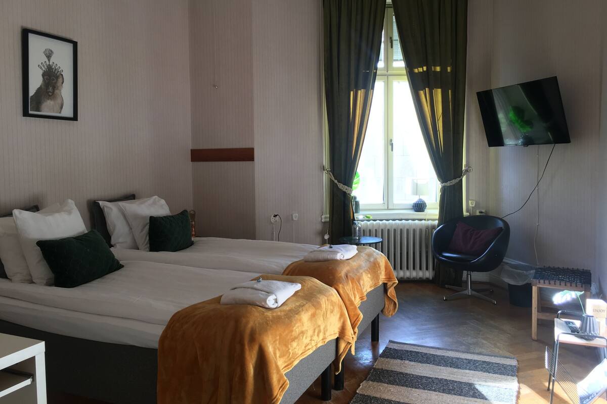 Hotellihuone, jossa on kaksi sänkyä, televisio, tuoli ja pieni pöytä, jossa on lamppu.