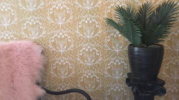 A corner with a patterned wallpaper, a potted plant, and a fluffy pink pillow on a chair.