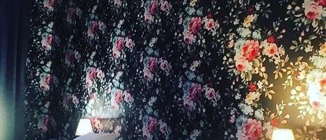 A hotel room with a bed, pillows, a bedside table with a lamp, and a floral wallpaper.