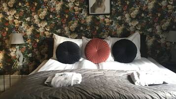 A neatly made bed with a floral wallpaper background, a framed picture above the bed, and a small stool in front.
