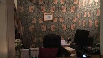 Et værelse med blomstret tapet, et skrivebord med en computer, en stol og et lille bord med en vase og en skål.