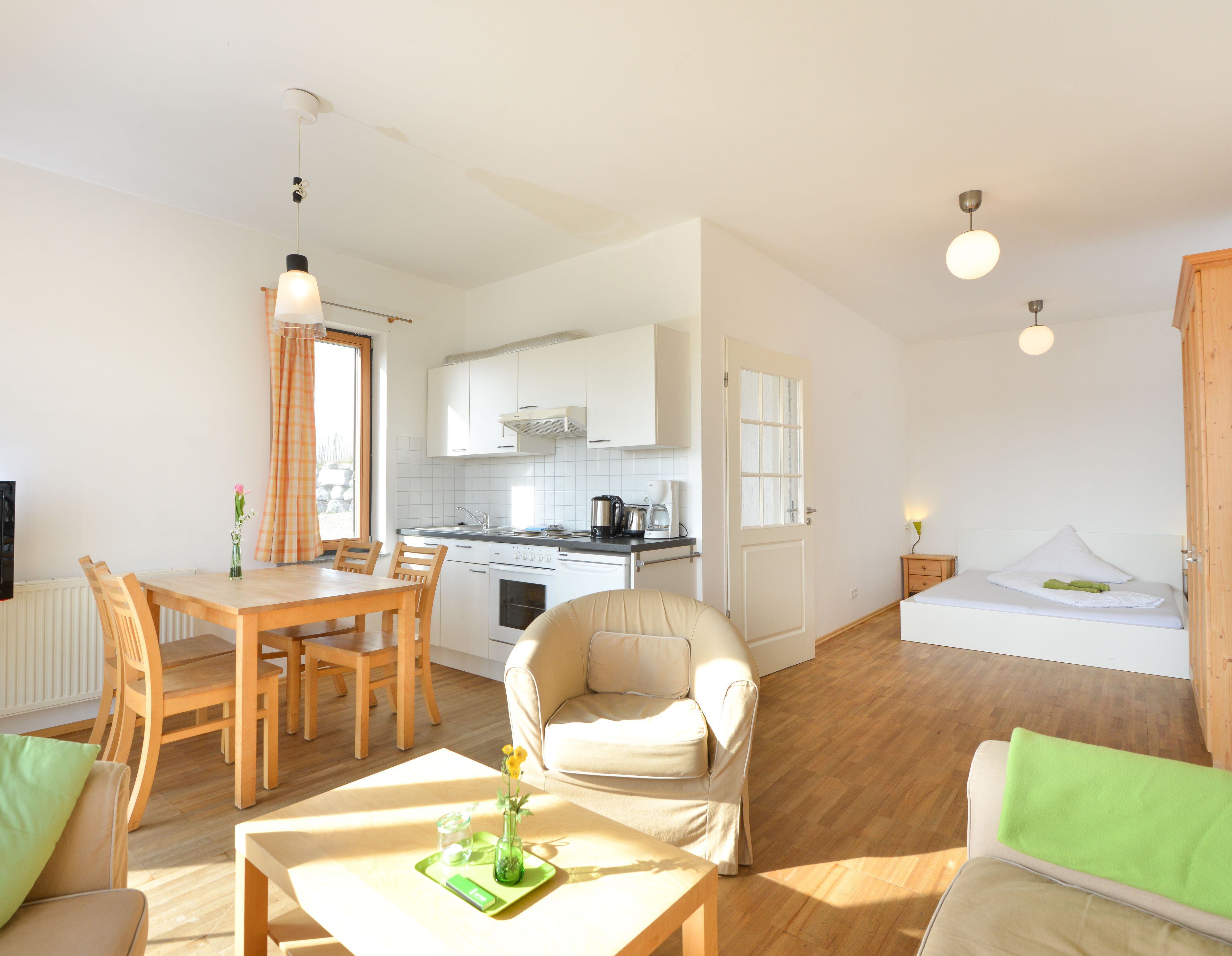 A modern, well-lit living room with a dining area, a kitchenette, and a bedroom with a bed and a nightstand.