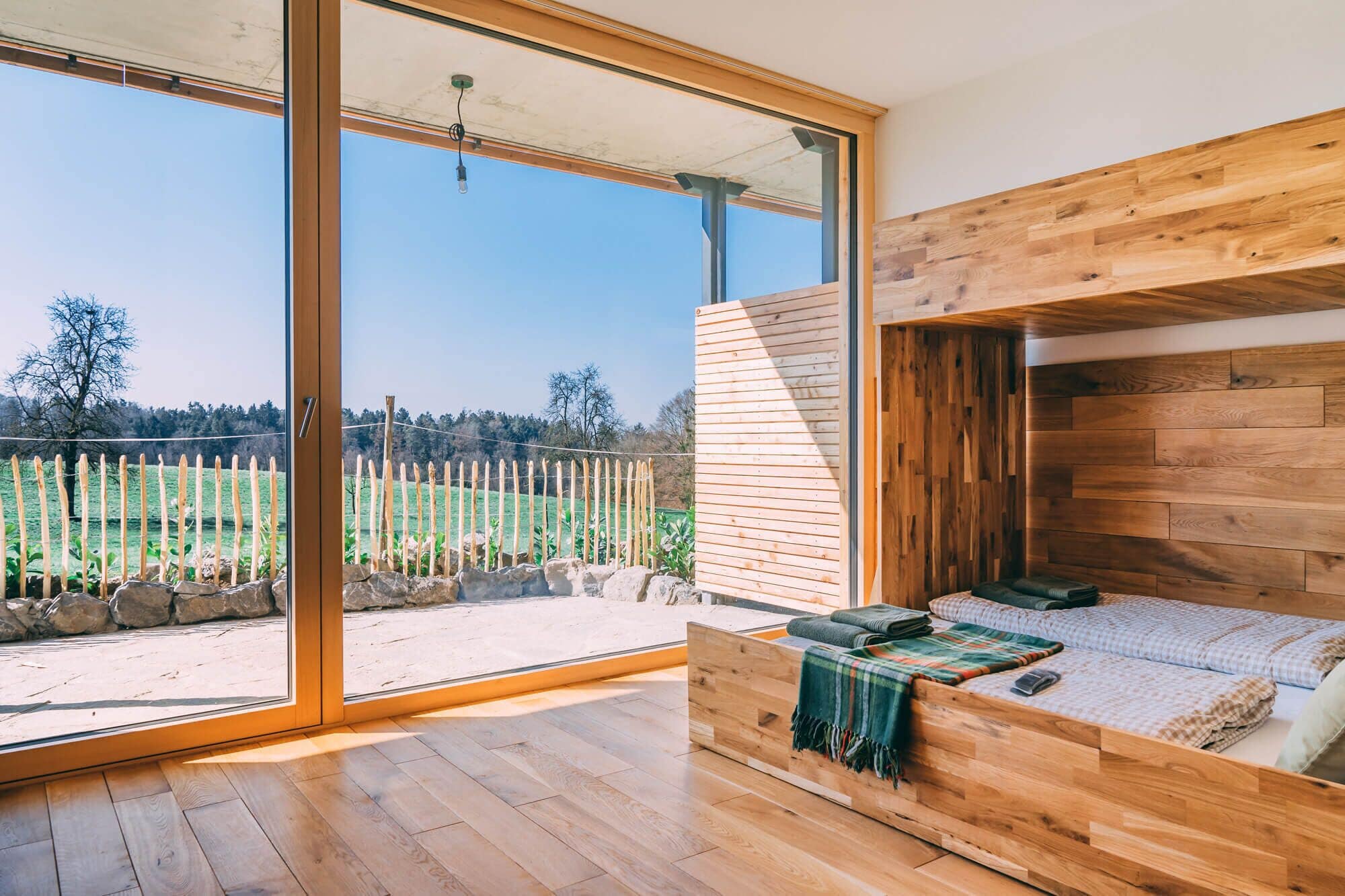 Ein modernes Schlafzimmer mit einem hölzernen Bett, einer großen Schiebeglastür, die auf einen Balkon führt, und Blick auf ein grünes Feld.