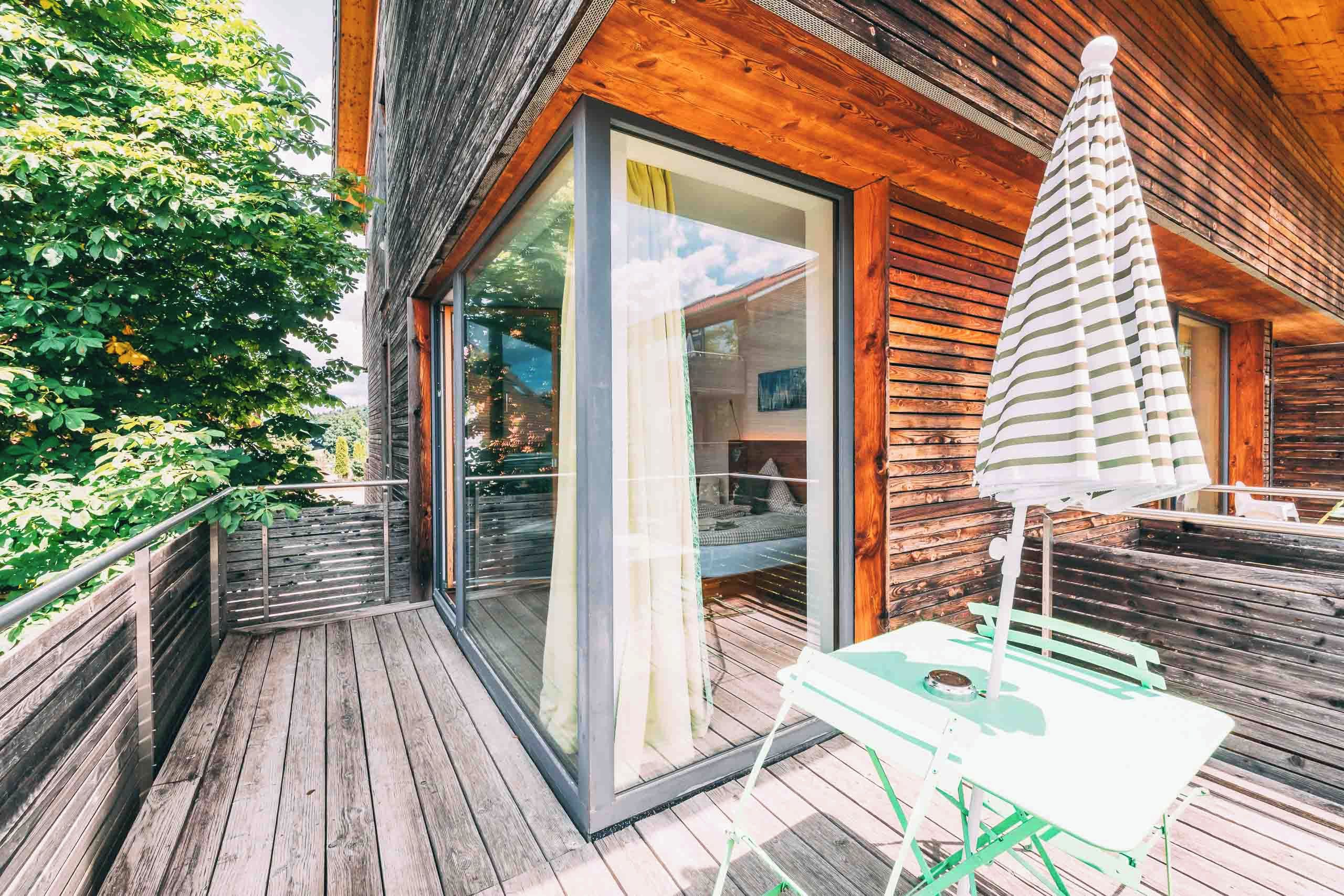 Eine HolzdTerrasse mit Glastür, die zu einem Raum führt, einem Tisch mit gestreiftem Sonnenschirm und Blick auf einen Garten.