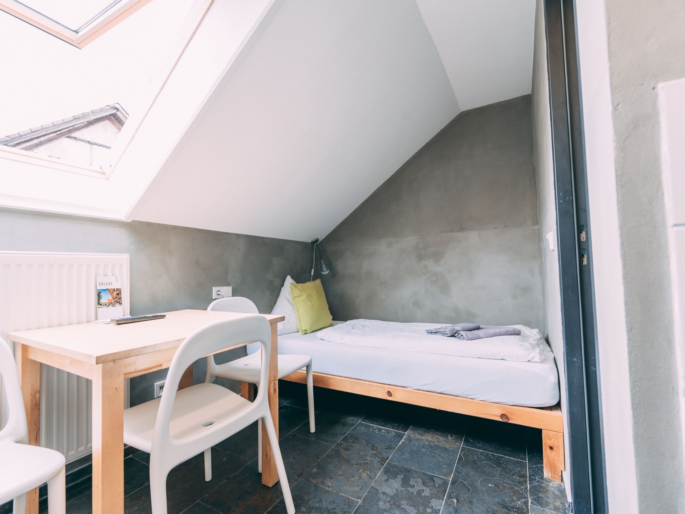 A small, modern room with a sloped ceiling, a single bed, a table with chairs, and a radiator.