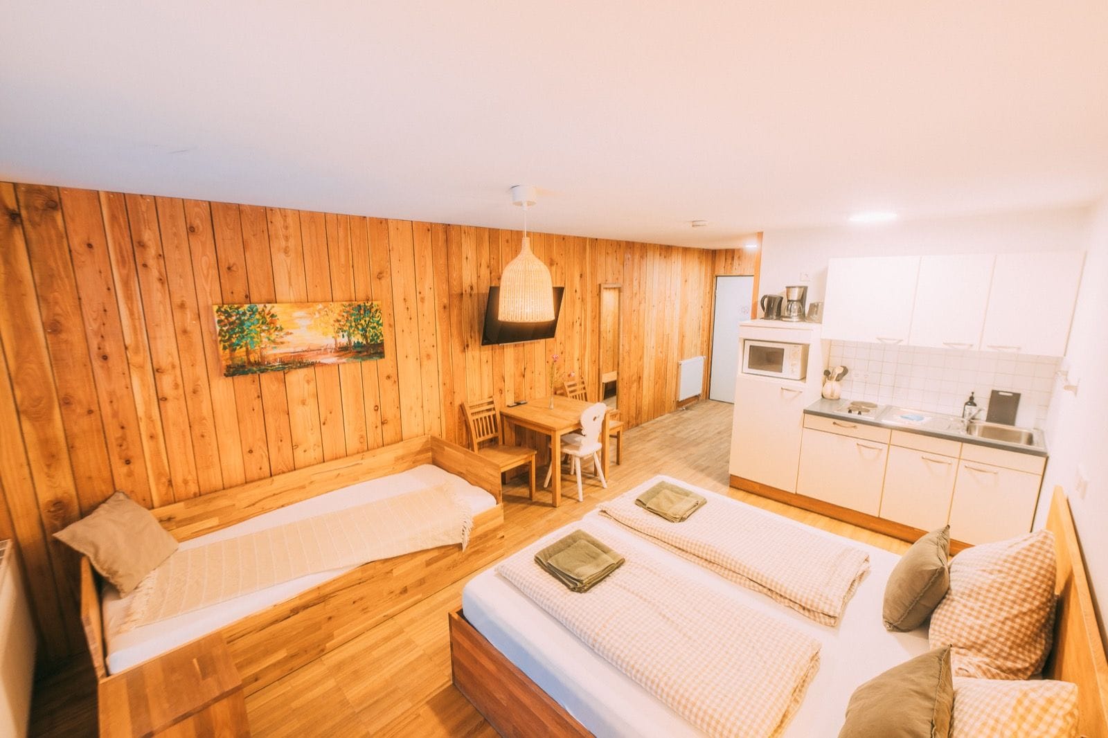 A room with two beds, a wooden wall, a dining table, and a kitchenette.