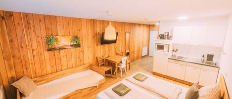 A room with two beds, a wooden wall, a dining table, and a kitchenette.