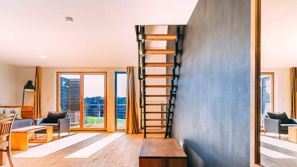 Ein modernes Wohnzimmer mit Holzboden, einer zentralen Holzbank, einer Treppe und großen Fenstern mit Blick ins Freie.