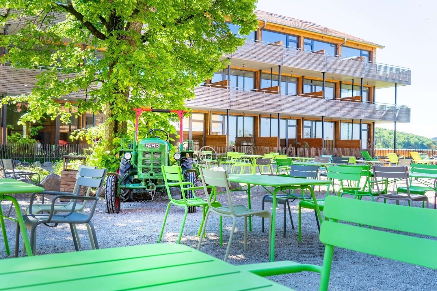An outdoor seating area with green tables and chairs, a tractor, and a modern building with large windows in the background.
