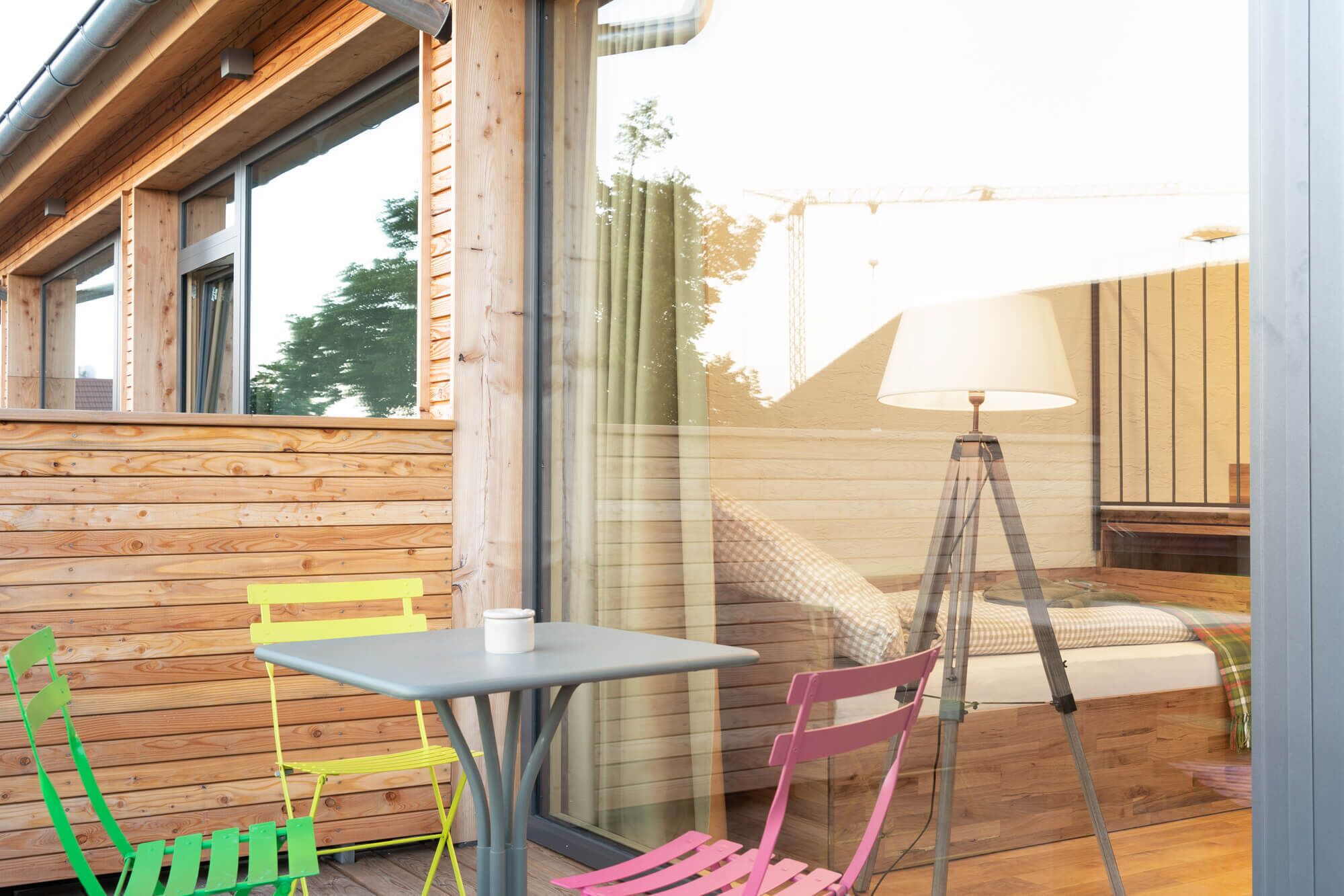 Een houten terras met een tafel, stoelen, een lamp en een bed met een geruit plaid.