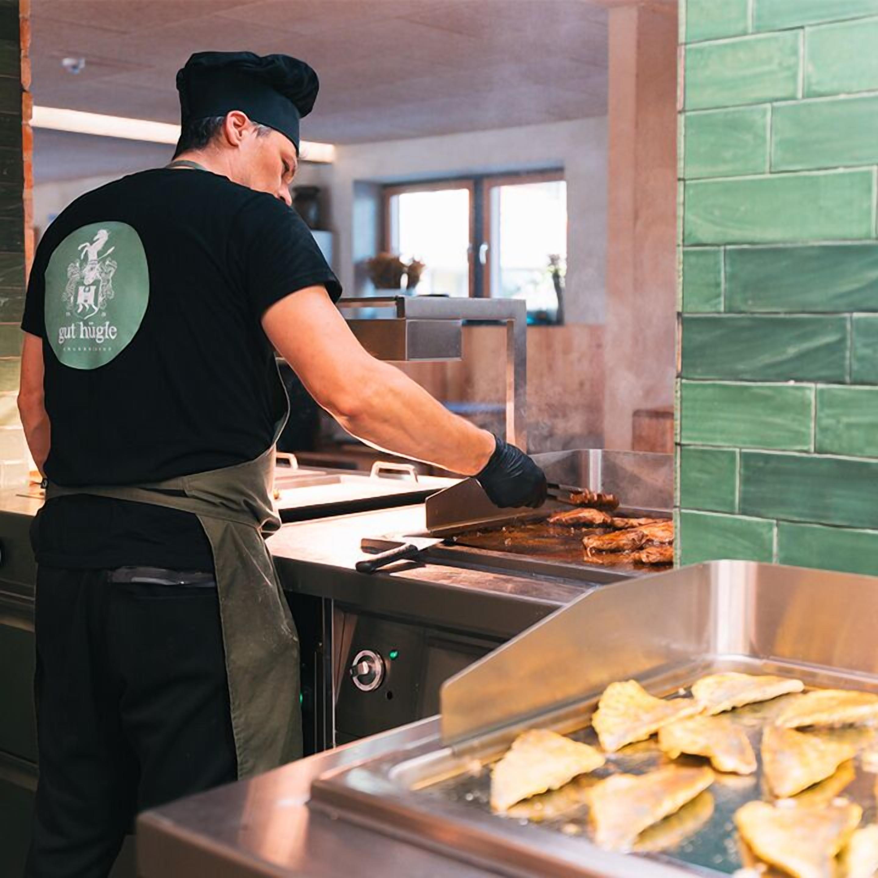 A chef cooking at a commercial kitchen with a green tiled wall and a stainless steel grill.