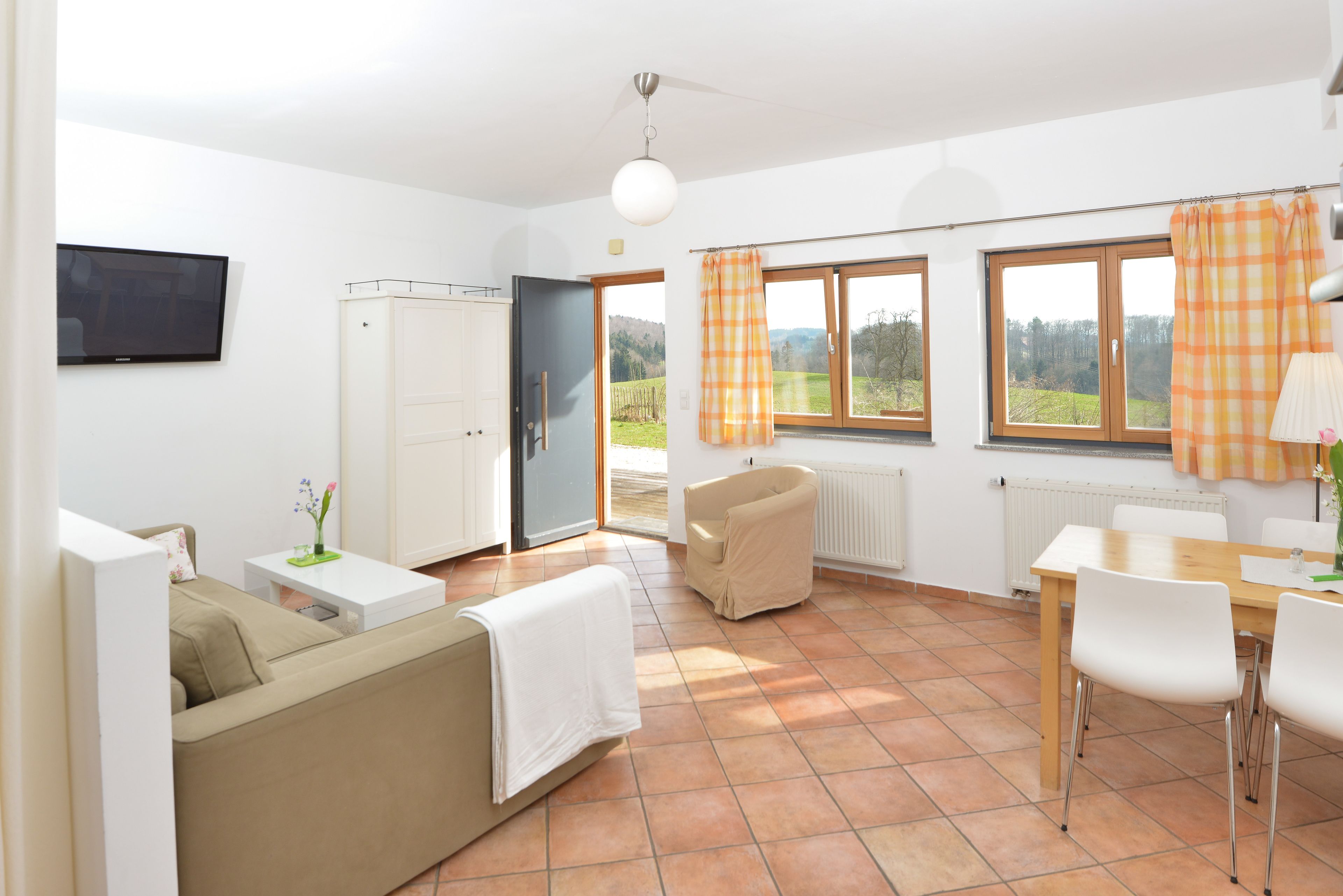 Een moderne woonkamer met een bank, een eettafel en stoelen. Er is een televisie aan de muur bevestigd, een raam met gordijnen en een radiator.