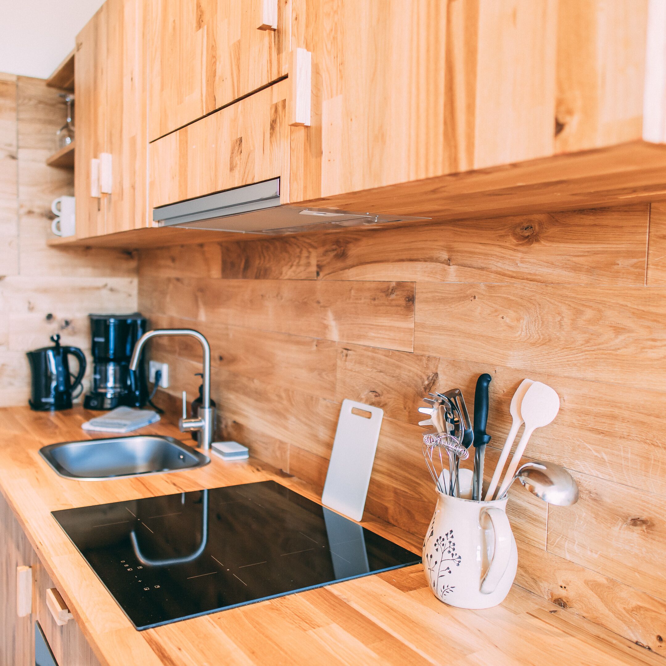 A modern kitchen with wooden cabinets, a stainless steel sink, a black induction cooktop, and various kitchen utensils.