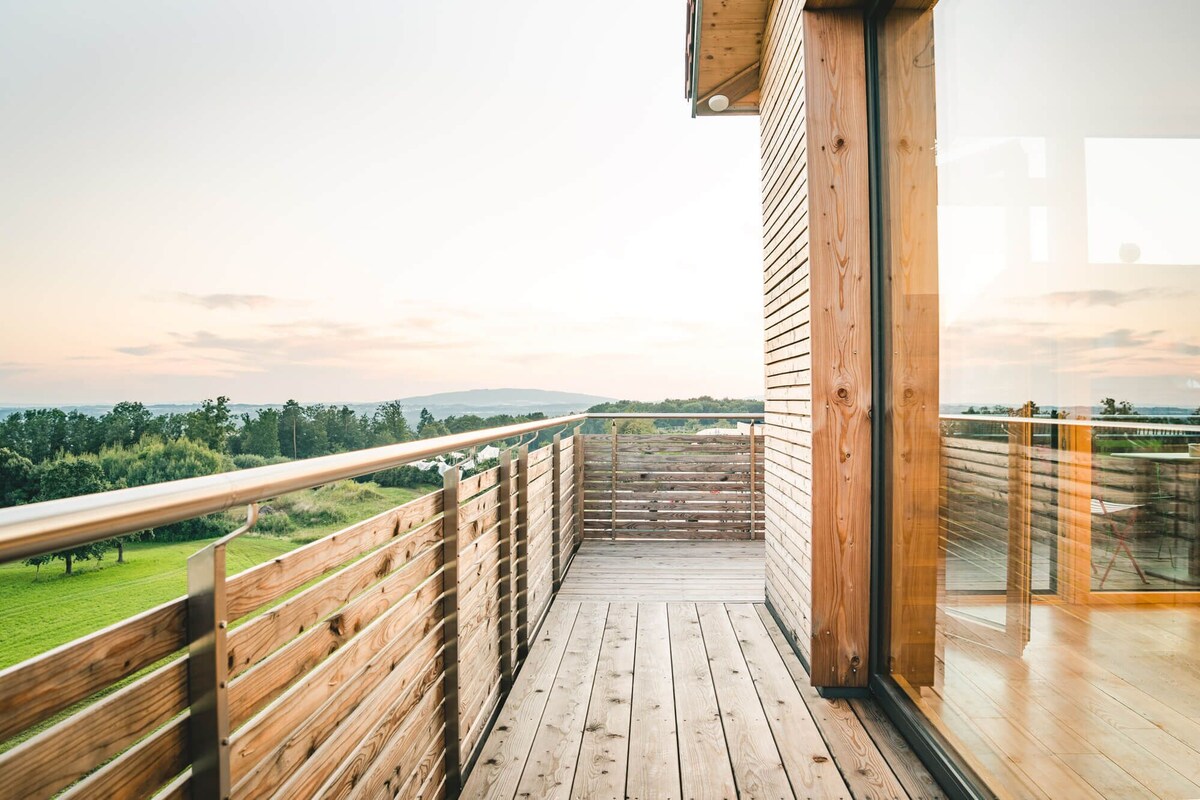 Una terraza de madera con barandilla metálica, que da a una puerta de vidrio con vistas a un paisaje verde y colinas distantes.