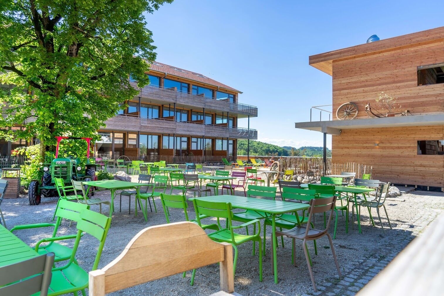 An outdoor seating area with green tables and chairs, a tractor, and a building with a wooden facade and large windows.
