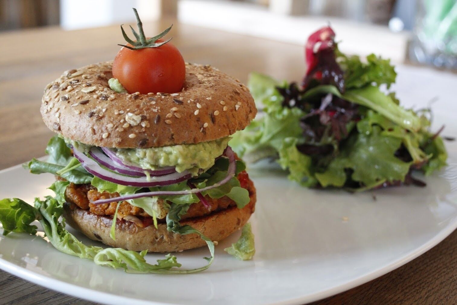 Een sandwich met een tomaat erop, omringd door groenten en een kleine salade op een wit bord.