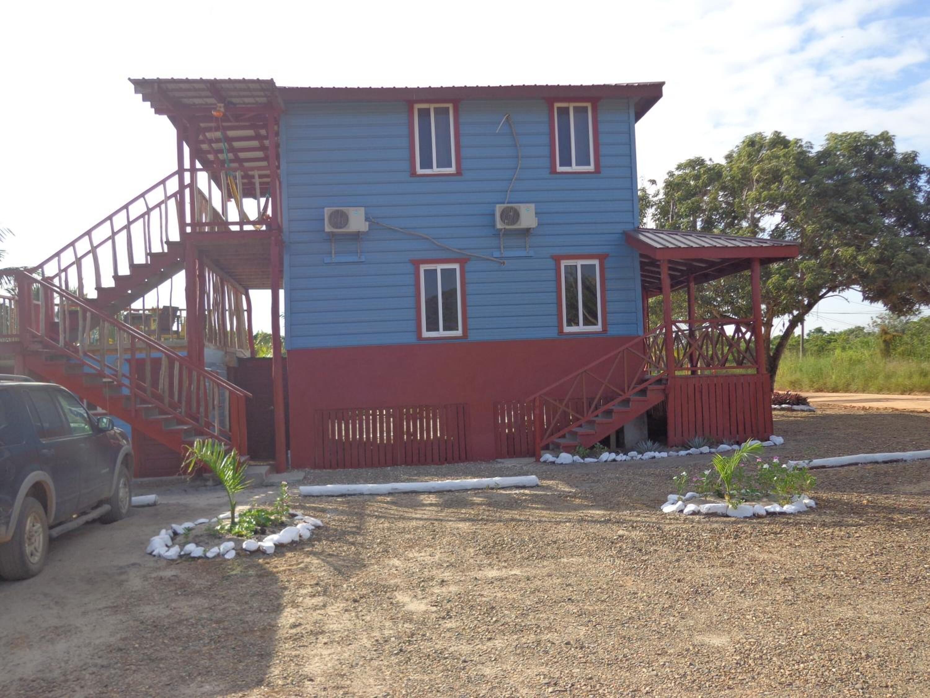 Un edificio de dos pisos con fachada azul y detalles rojos, que incluye una escalera y un porche cubierto.