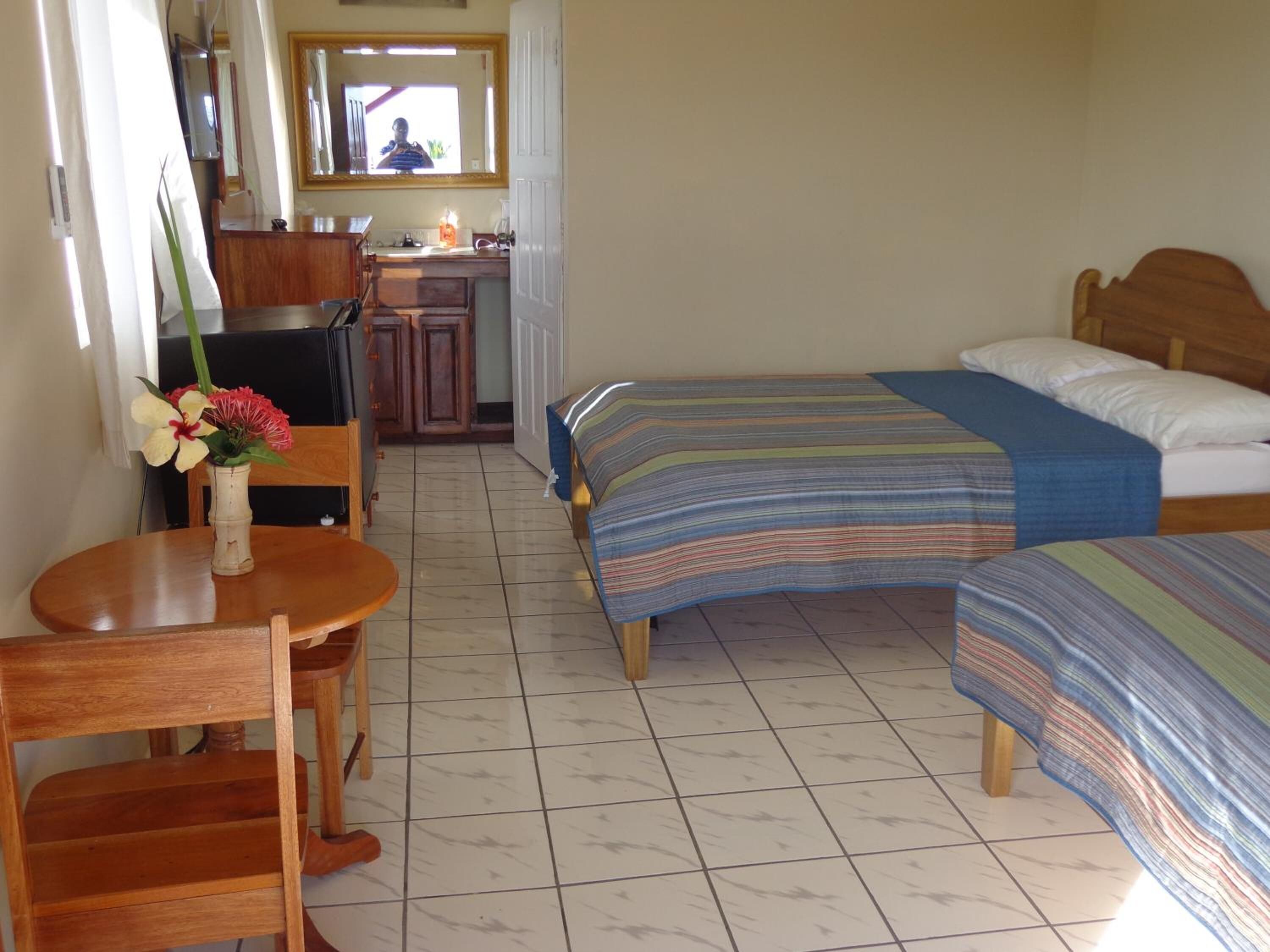Ein Zimmer mit zwei Betten, einem Holztisch, einem Stuhl, einer kleinen Küchenzeile und einem durchgegangene Tür erkennbaren Badezimmer.
