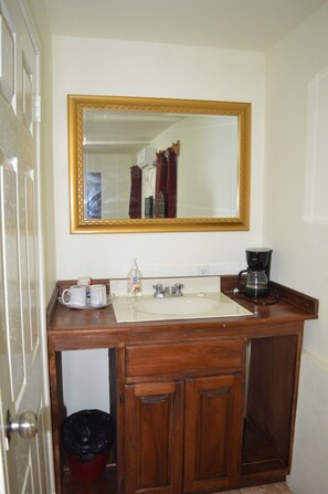A bathroom with a wooden vanity, a mirror with a gold frame, a sink, a coffee maker, and a trash bin.