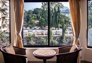 Quarto com uma grande janela, duas poltronas estampadas, uma pequena mesa redonda e vista para um estacionamento e edifícios.