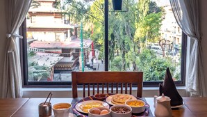 Una mesa de comedor con alimentos para el desayuno, vista de una construcción tradicional y una ventana con cortinas.