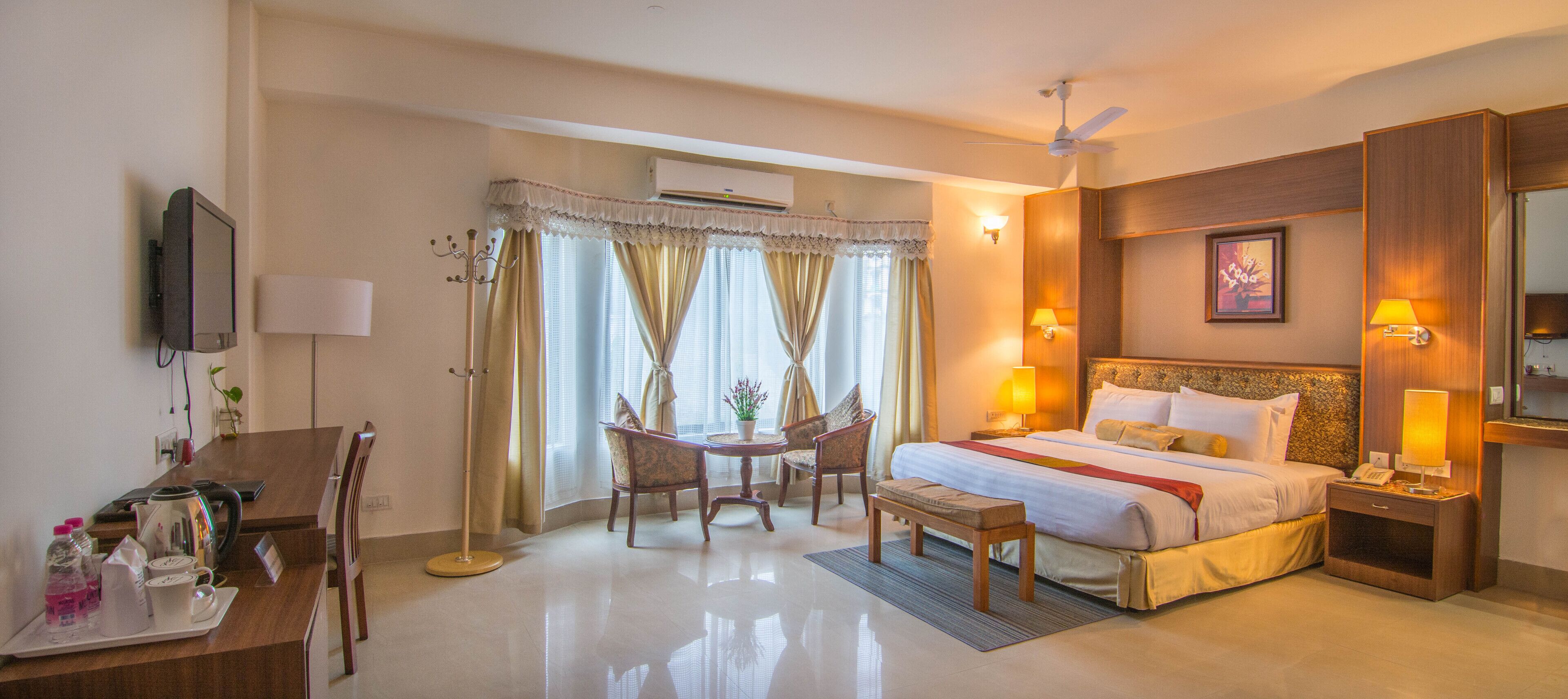 A hotel room with a bed, bedside tables, a desk with a chair, a small table with chairs, and a large window with curtains.