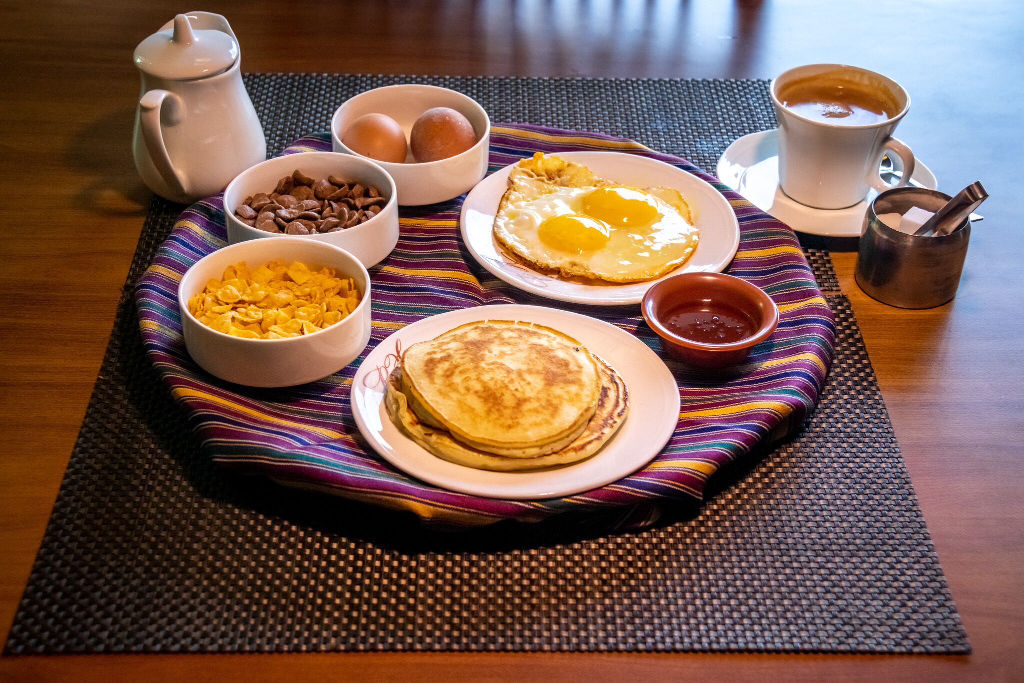 Een ontbijttafel met pannenkoeken, eieren en koffie op een geweven onderzetter.