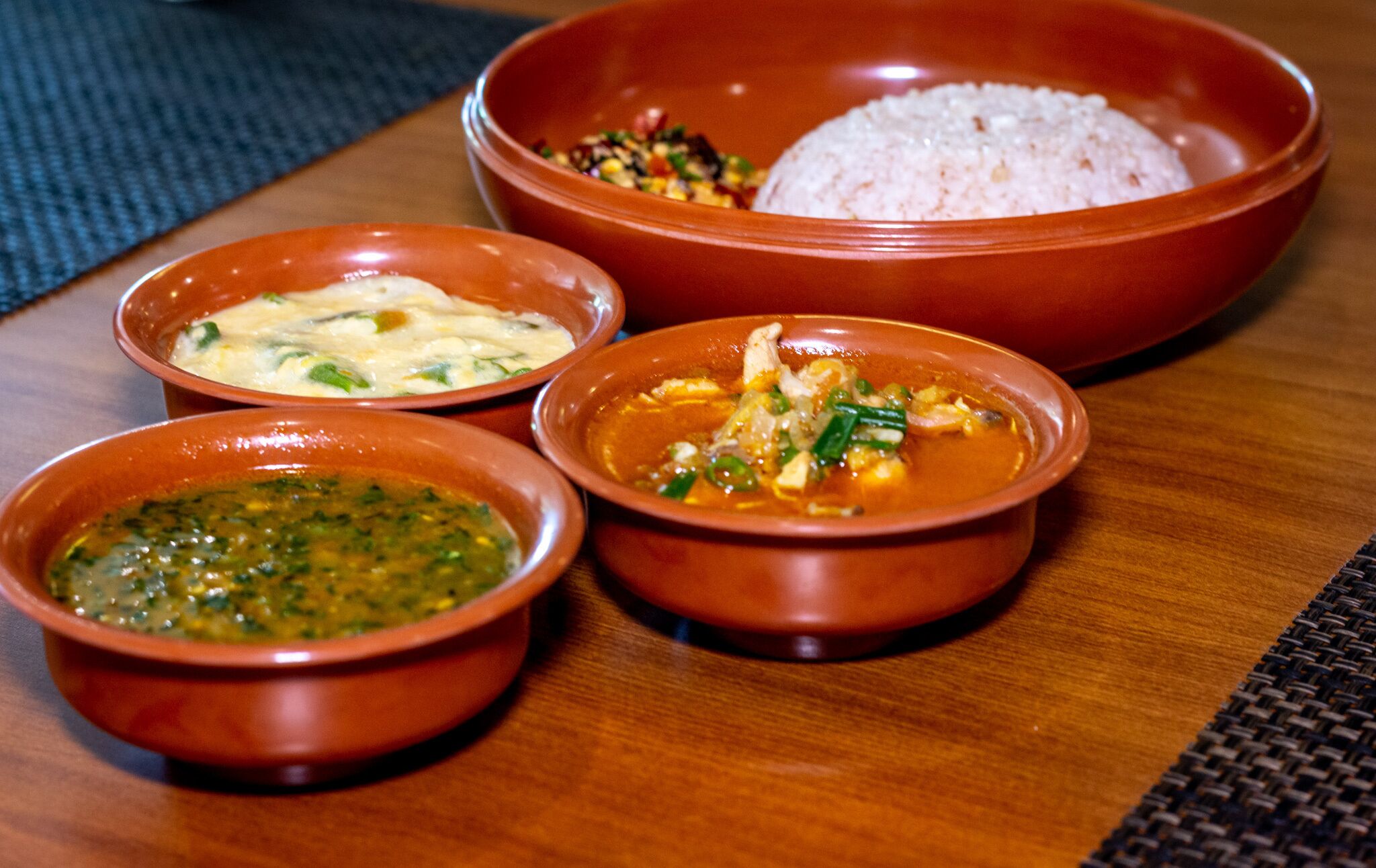 Una mesa de madera con cuatro cuencos que contienen diferentes platos, incluyendo arroz y una salsa verde.