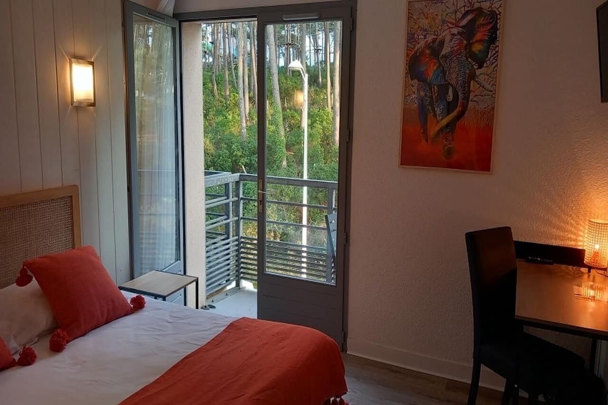 A bedroom with a bed, a desk, and a balcony with a view of trees.
