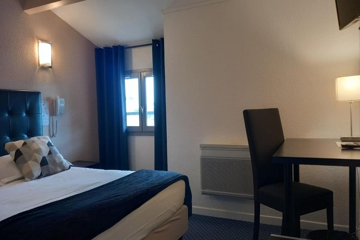 A hotel room with a bed, a desk, a chair, a lamp, and a window with curtains.
