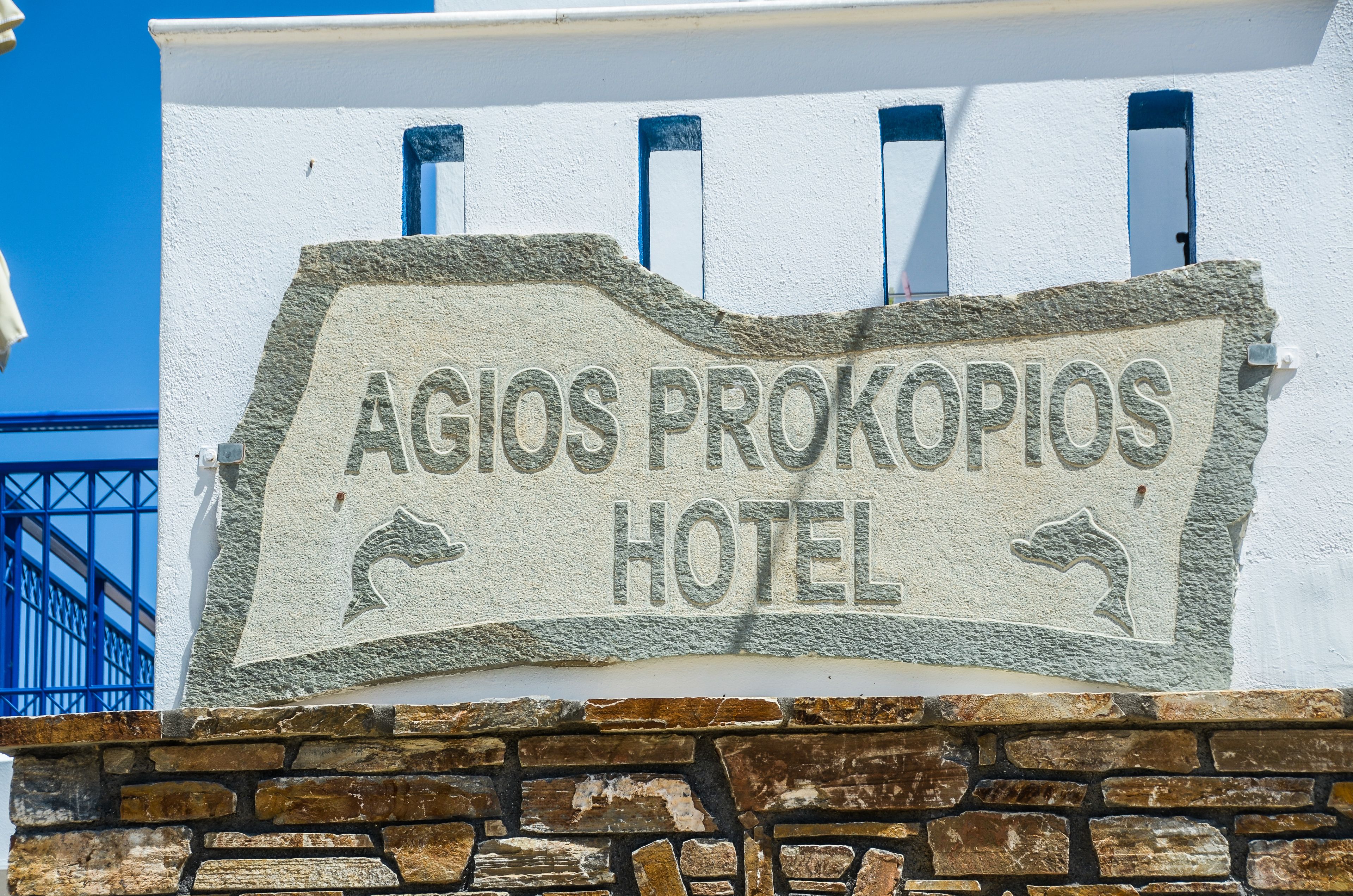 A stone sign for the Agios Prokopios Hotel with a stone wall and blue window frames.