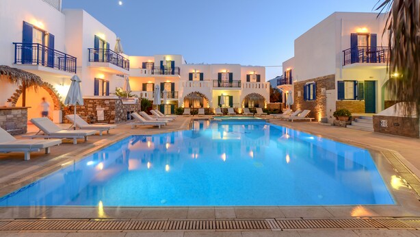 Una piscina rodeada de edificios blancos con balcones, sillas de descanso y sombrillas.