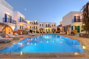 Una piscina rodeada de edificios blancos con balcones, sillas de descanso y sombrillas.