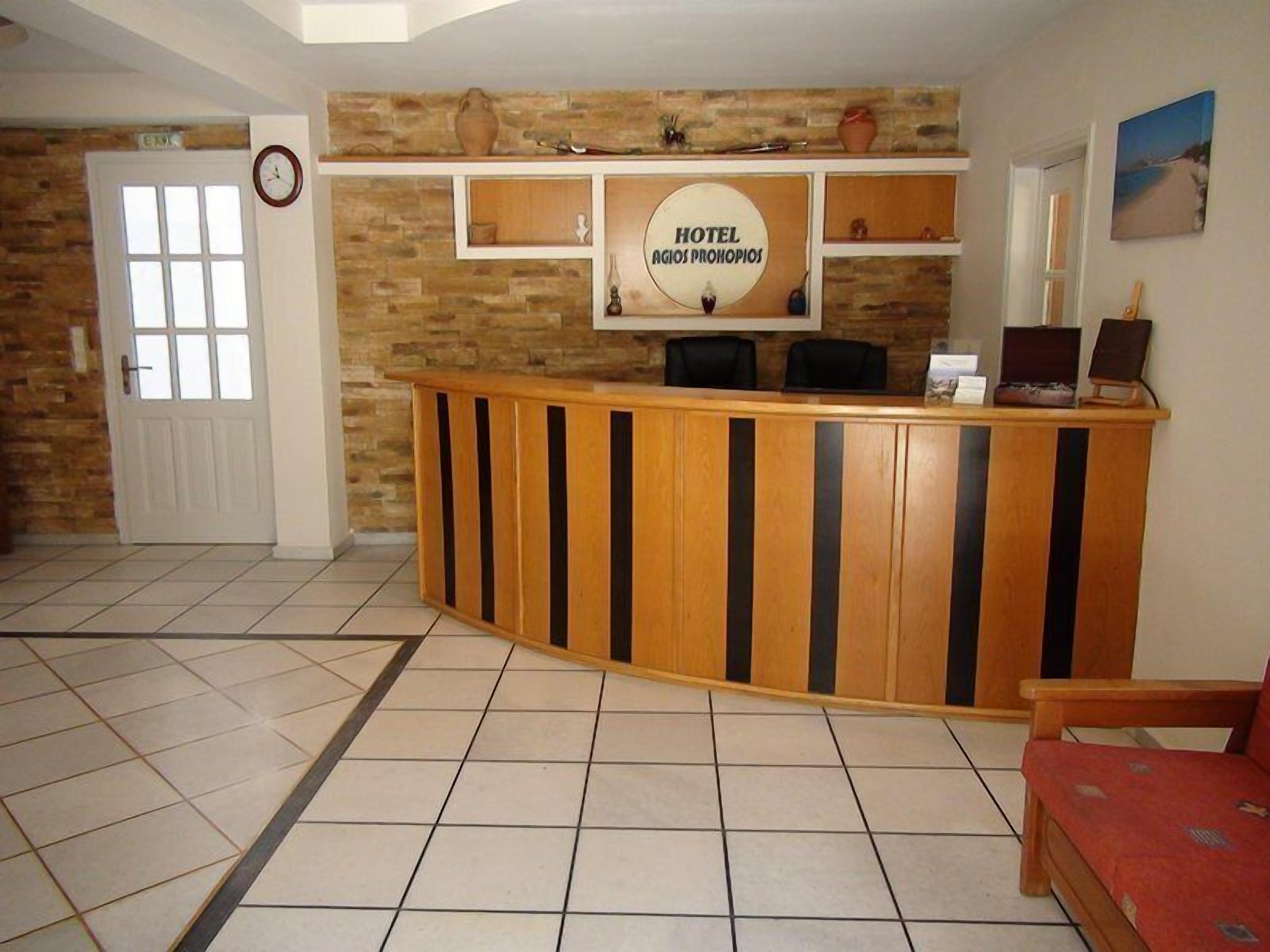 A reception area with a wooden reception desk, a clock, and a sign that reads "HOTEL AGIOS PROHOPIOS".