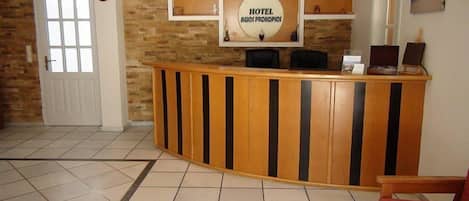 Un espace d’accueil avec un comptoir de réception en bois, une horloge et une enseigne indiquant « HÔTEL AGIOS PROHOPIOS ».