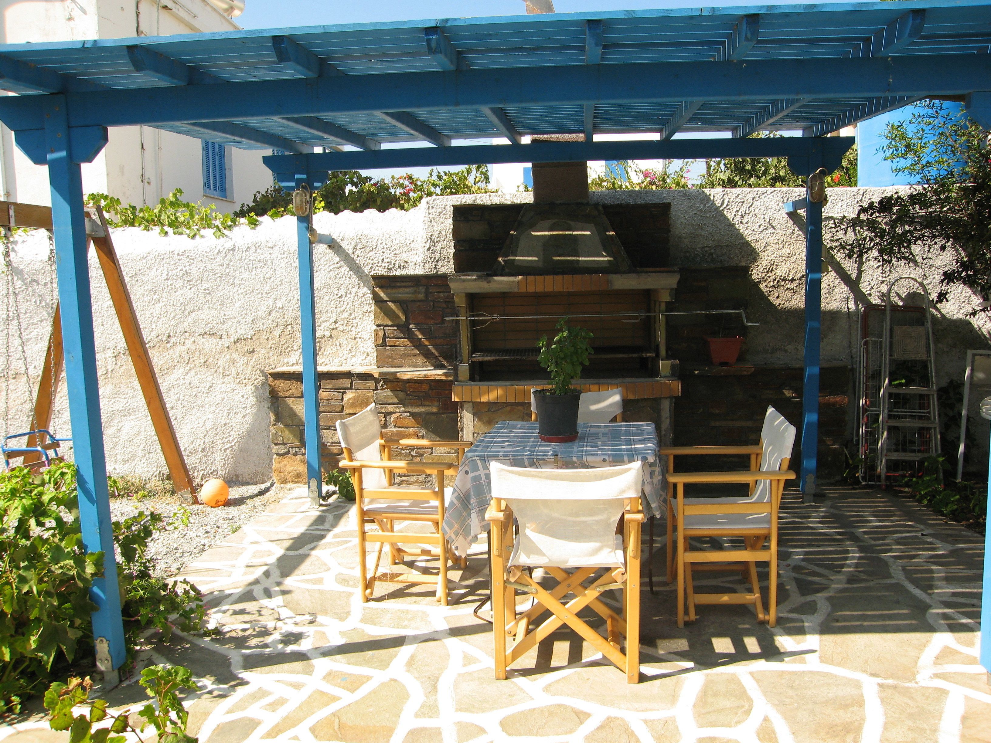 An outdoor patio with a stone floor, wooden chairs, a table, and a blue pergola.