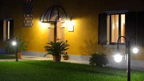 A building with a yellow facade, a brick entrance, and a well-lit outdoor area with a pathway, potted plants, and a small garden. - Agriturismo Bacche di Bosco (Verona)