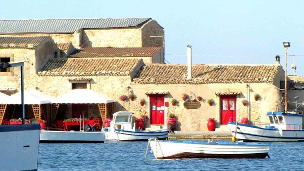 A coastal building with a tiled roof, a small dock with boats, and outdoor seating with umbrellas. - Casa Vacanza Ondablu (Ispica)