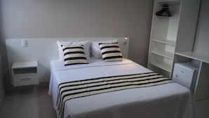 A neatly made bed with striped bedding, a white nightstand with a drawer, and a white wardrobe with shelves. - Ventura Hotel (Barreiras)