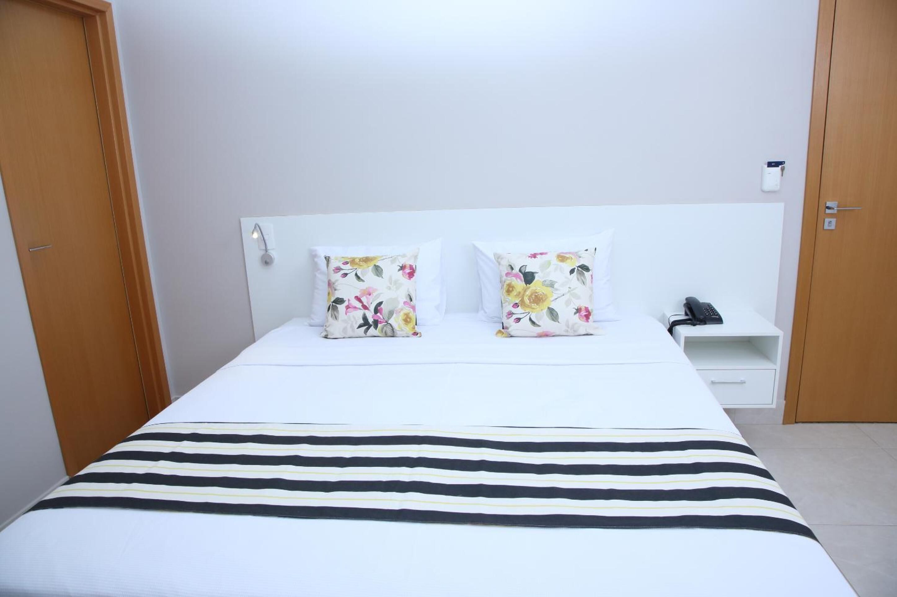 Habitación de hotel con una cama, dos almohadones florales, una mesita de noche con un teléfono y una colcha a rayas.