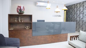 A reception desk with a modern design, featuring a glass top and wooden shelves. There are two pendant lights above the desk, and a wall-mounted air conditioning unit is visible. - Ventura Hotel (Barreiras)