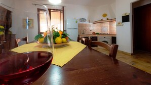 Ein Essbereich mit einem Holztisch, Stühlen und einer Glasschale mit Früchten. Im Hintergrund ist eine Küche mit gefliestem Fliesenspiegel und Geräten erkennbar.