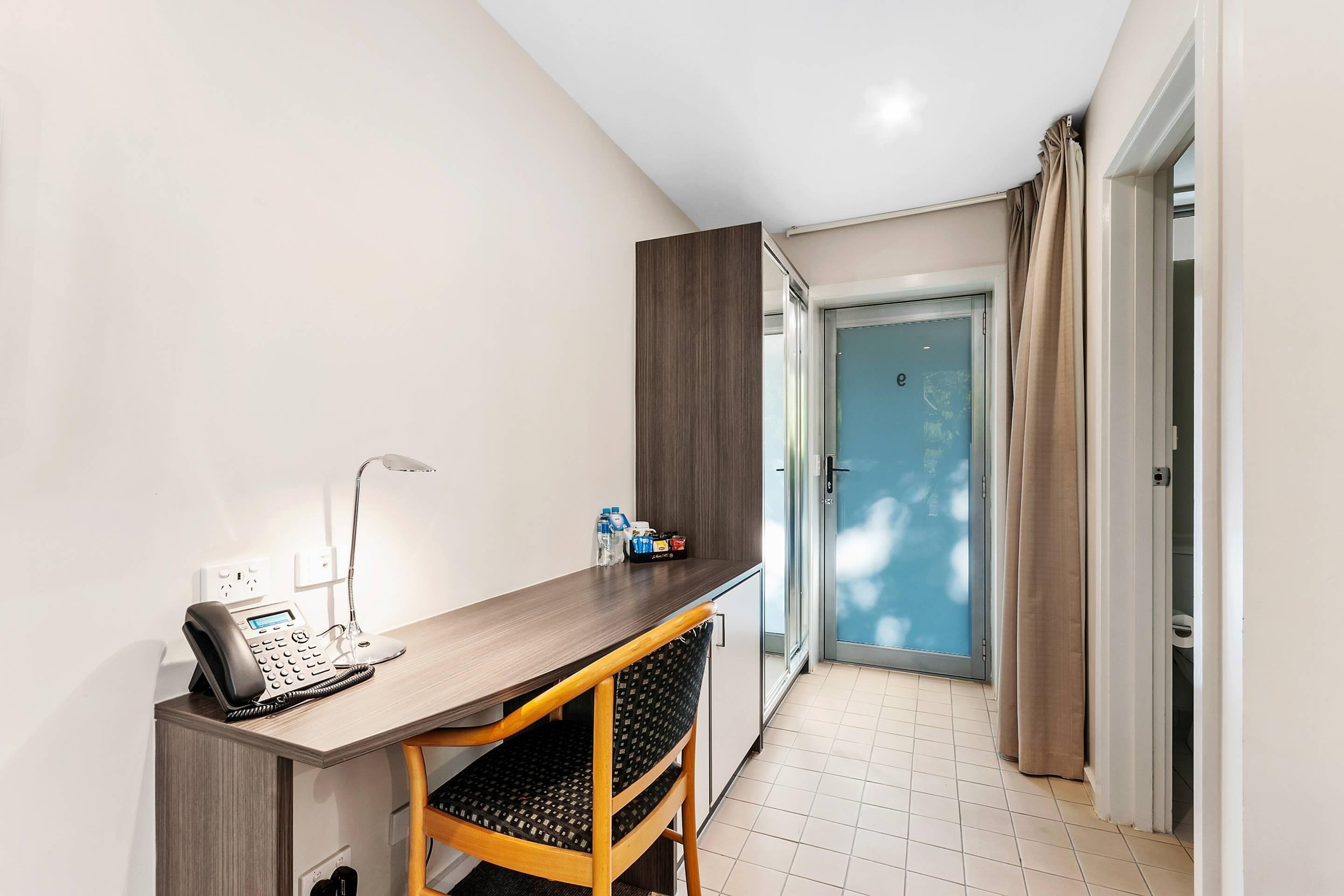 A hotel room with a desk, chair, telephone, lamp, and a door leading to a balcony.