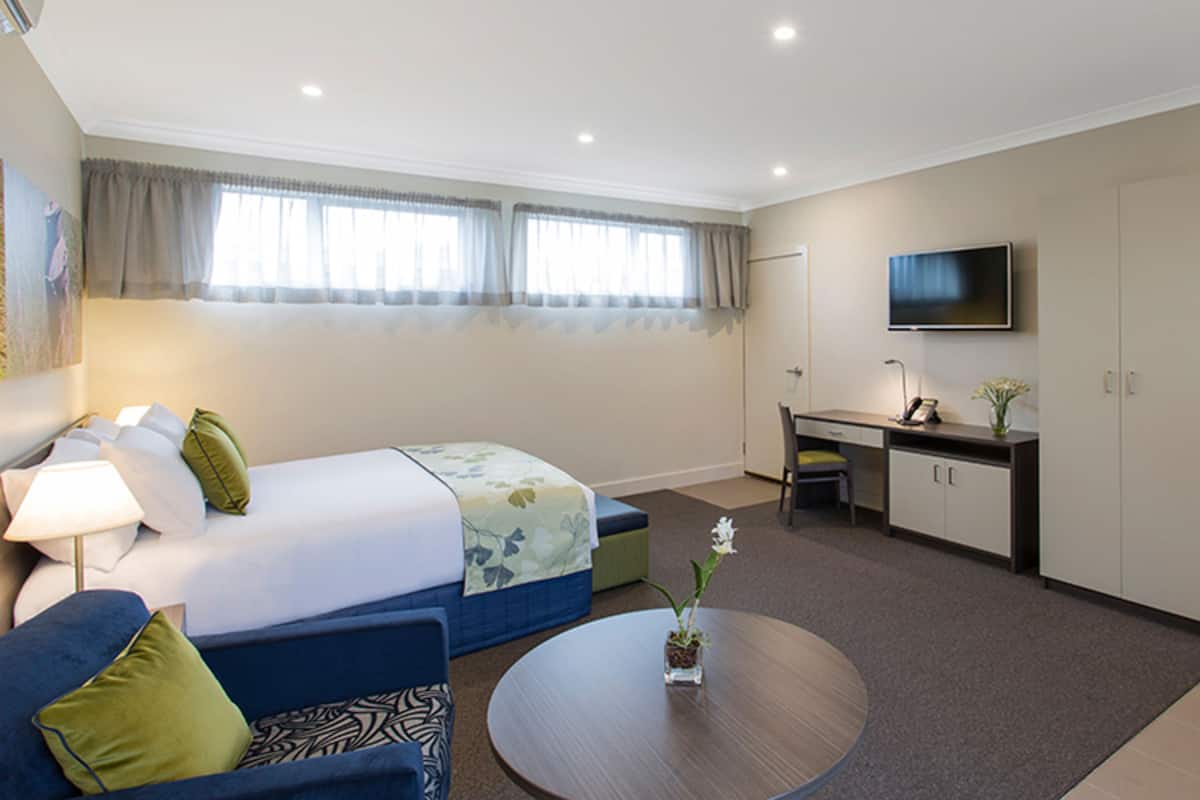 A hotel room with a bed, a desk, a chair, a TV, and a small table with a vase.