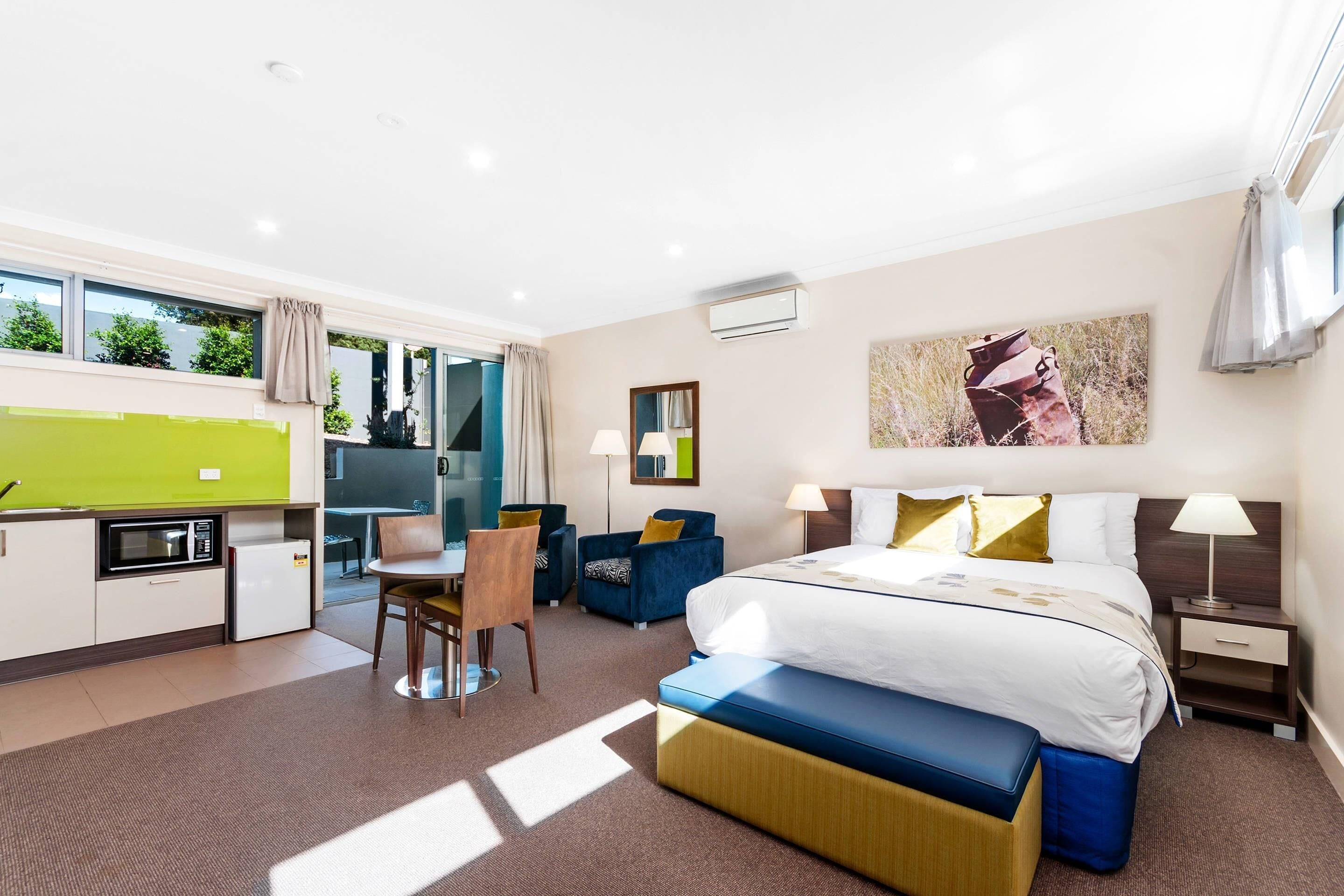 A modern hotel room with a bed, bedside tables, a desk, a chair, a small kitchenette, and a large window.