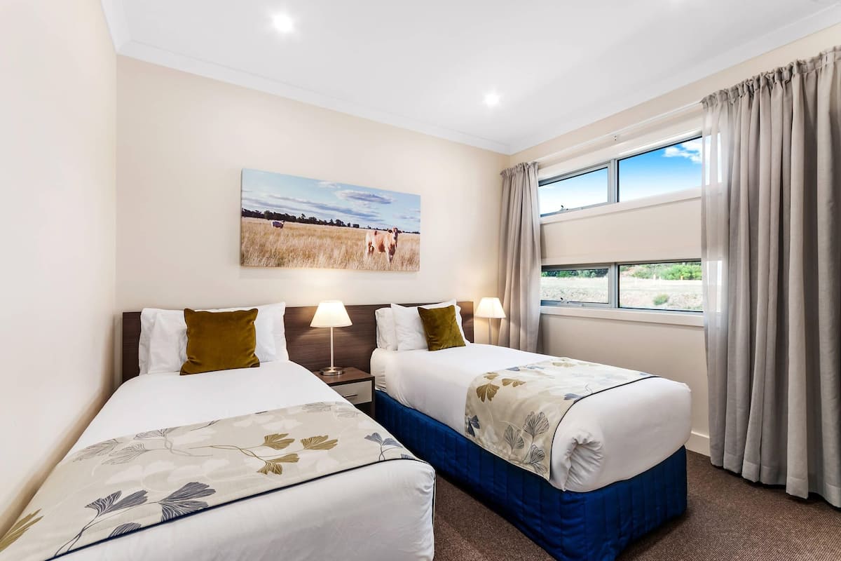 A hotel room with two beds, a painting of a field, a window with curtains, and a nightstand with lamps.