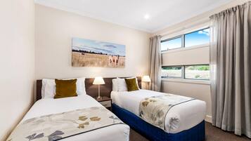 A hotel room with two beds, a painting of a field, a window with curtains, and a nightstand with lamps.
