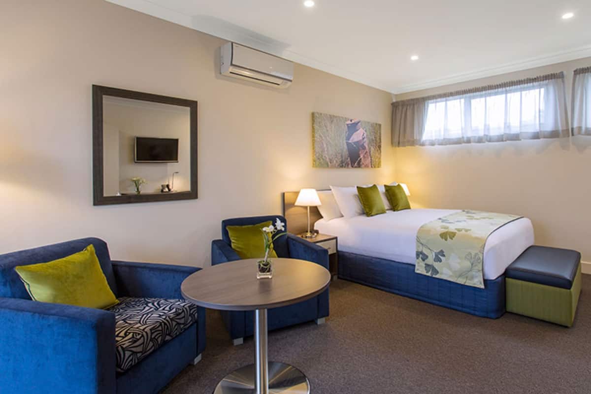 A hotel room with a bed, two blue armchairs, a round table, a TV, and an air conditioning unit.