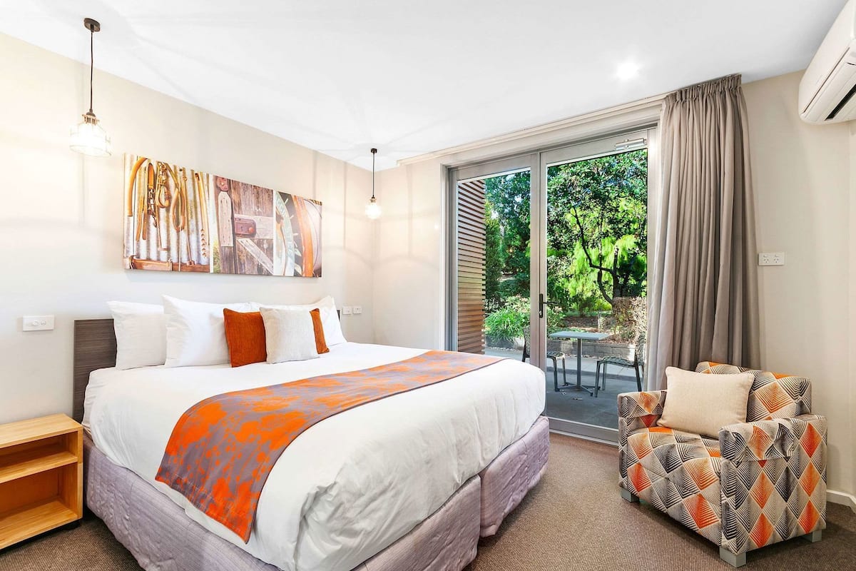 A hotel room with a large bed, a seating area with patterned cushions, a dining table outside, and a sliding glass door leading to a garden.